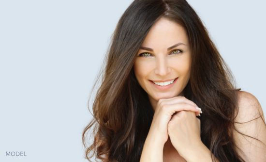 Brunette (MODEL) smiling at camera wth her hands clasped under her chin.